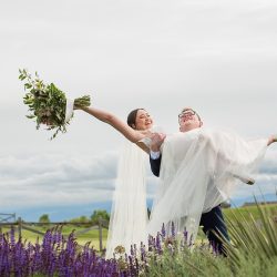 smiling bride holding her bouquet of flowers above her as groom lifts her in front of a golf course
